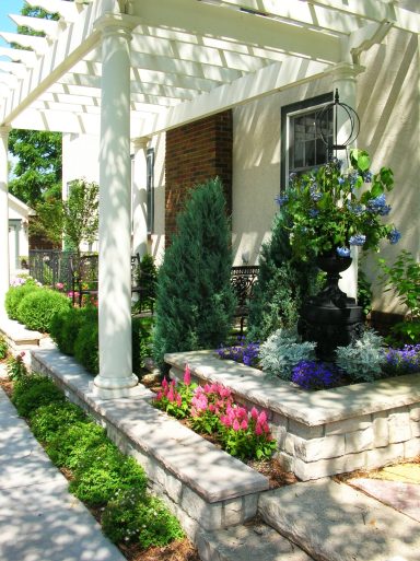 Lush garden with colorful flowers, greenery, and a decorative fountain beside a walkway.