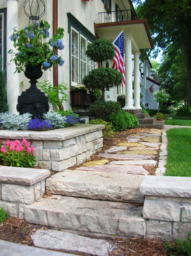 Stone steps lead to a house with vibrant flowers and an American flag in the background.