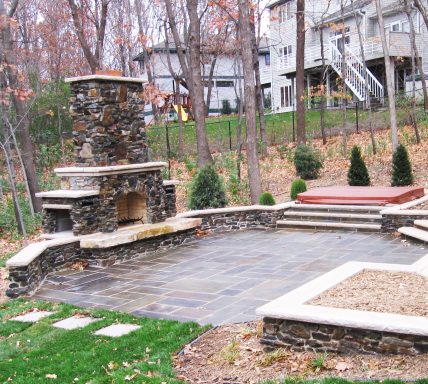 Stone outdoor fireplace with seating area and steps in a wooded backyard.