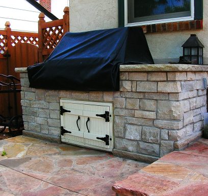 Stone outdoor grill with a black cover, set on a patio beside a wooden fence.