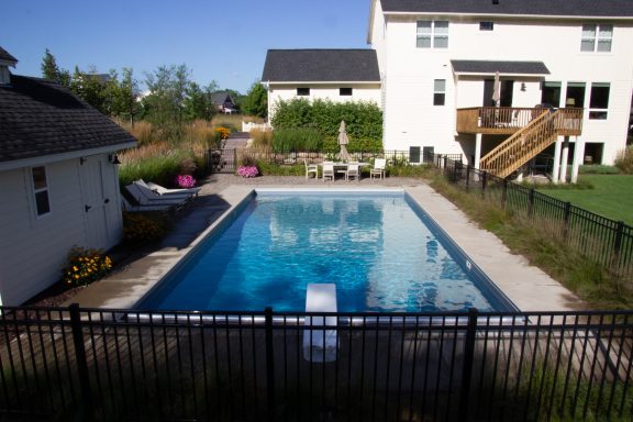 Backyard pool surrounded by a fence, grass, and patio furniture on a sunny day.