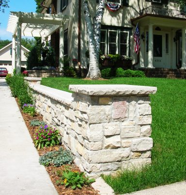 Stone wall with a rope design bordering a green lawn and flower beds near a house.