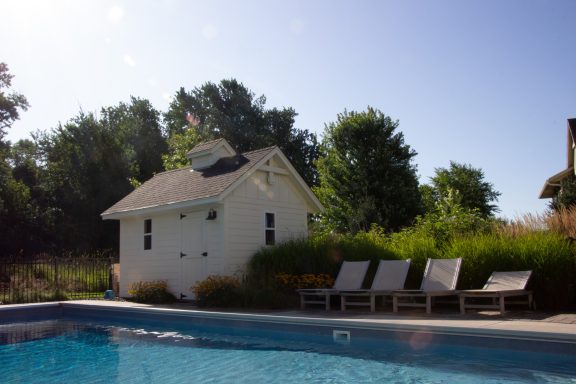 A quiet poolside scene featuring a small white house and lounge chairs surrounded by greenery.