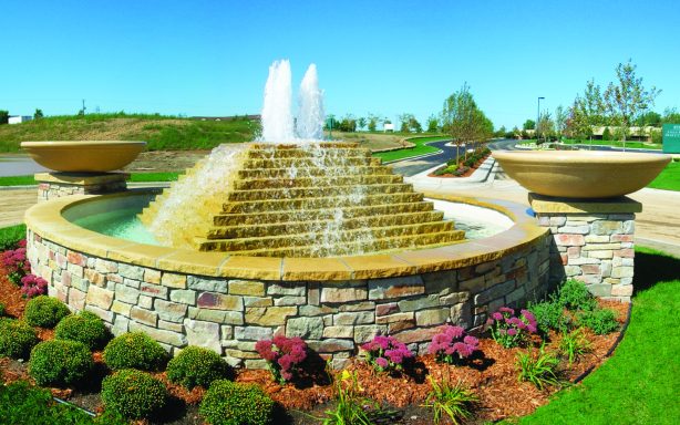 Tiered stone fountain with water jets surrounded by flowers and grass.