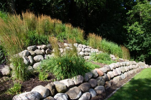A landscaped stone wall with decorative plants and grasses on a sloped garden.