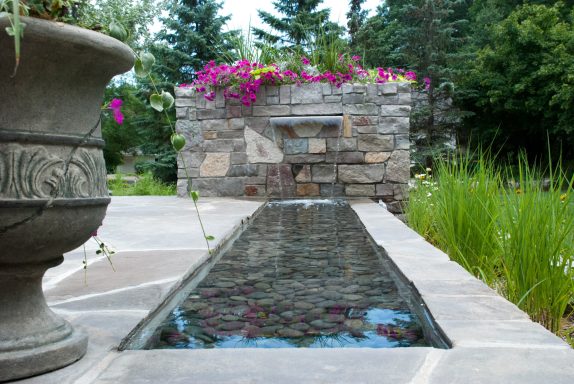 A stone water feature with flowers above, surrounded by greenery.