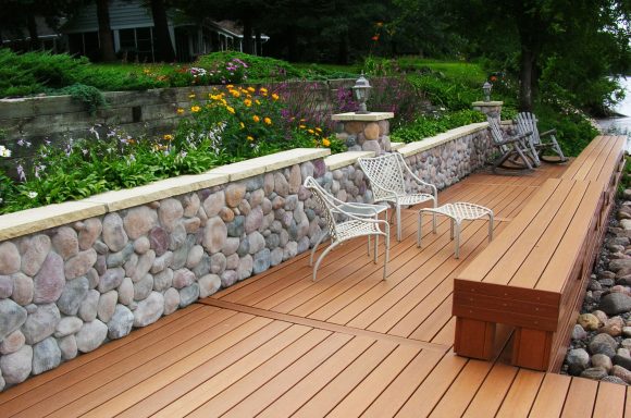Wooden deck with stone wall and chairs surrounded by colorful flowers and greenery.