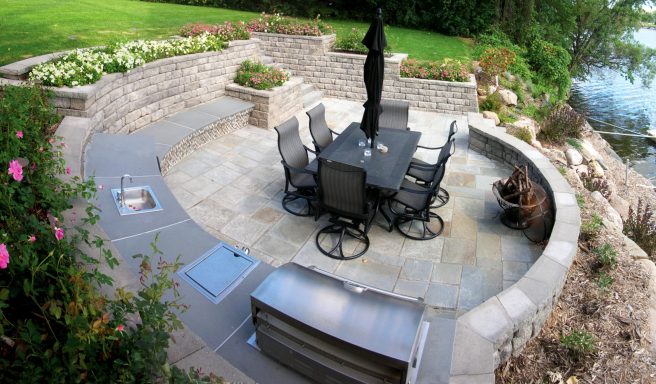 Patio area with dining set, umbrella, and landscaped garden by a body of water.