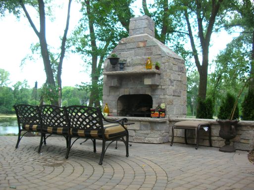 Outdoor stone fireplace with seating area and lush greenery nearby.