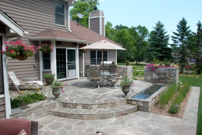 Patio area with stone flooring, dining set, umbrella, and planters under a blue sky.